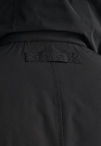 Sort jakke med en glat, mat finish og et syet GANT-mærke på bagsiden. Designet inkluderer krydssting-detaljer.