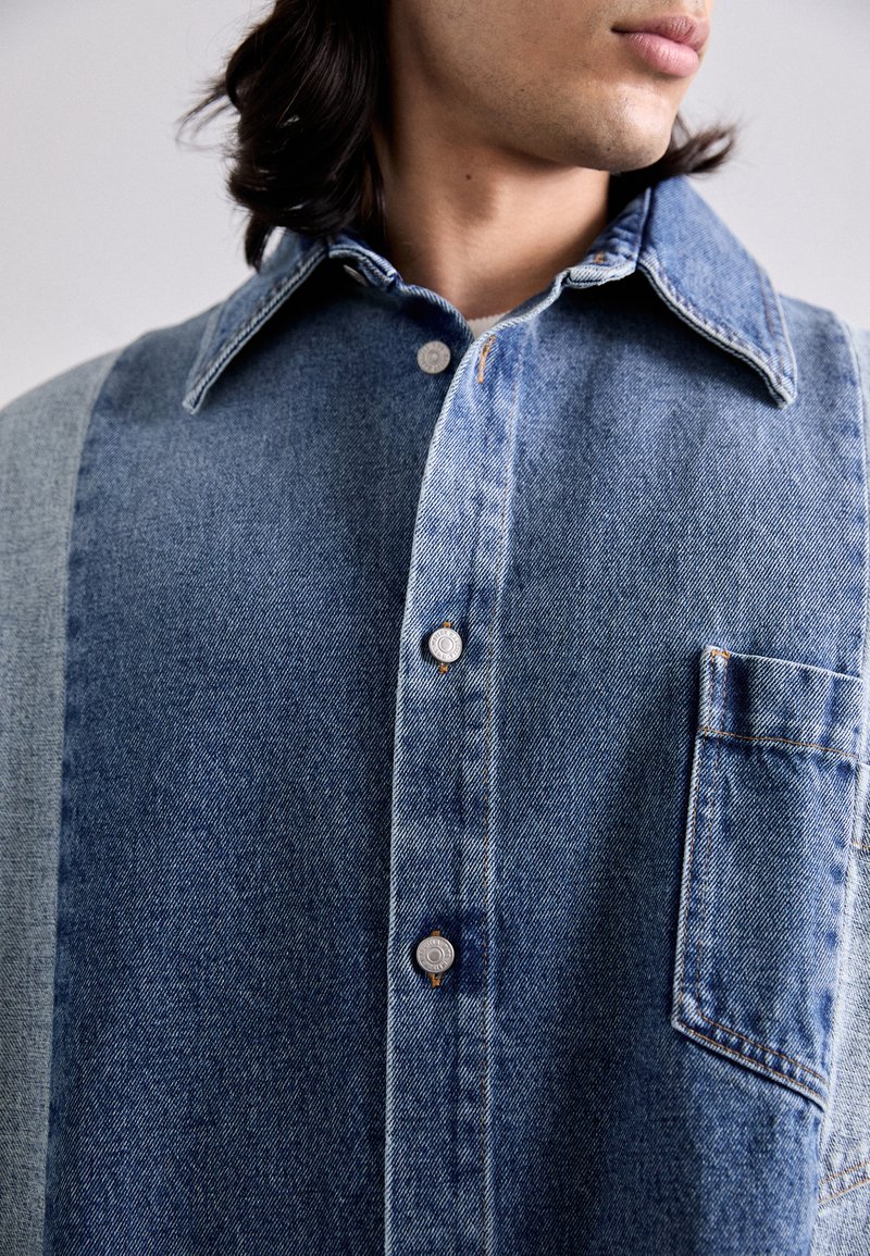 Camicia in denim con diverse tonalità di blu, colletto classico, chiusura con bottoni frontale e taschino singolo sul petto, che mette in mostra un tessuto testurizzato e dettagli di cucitura.