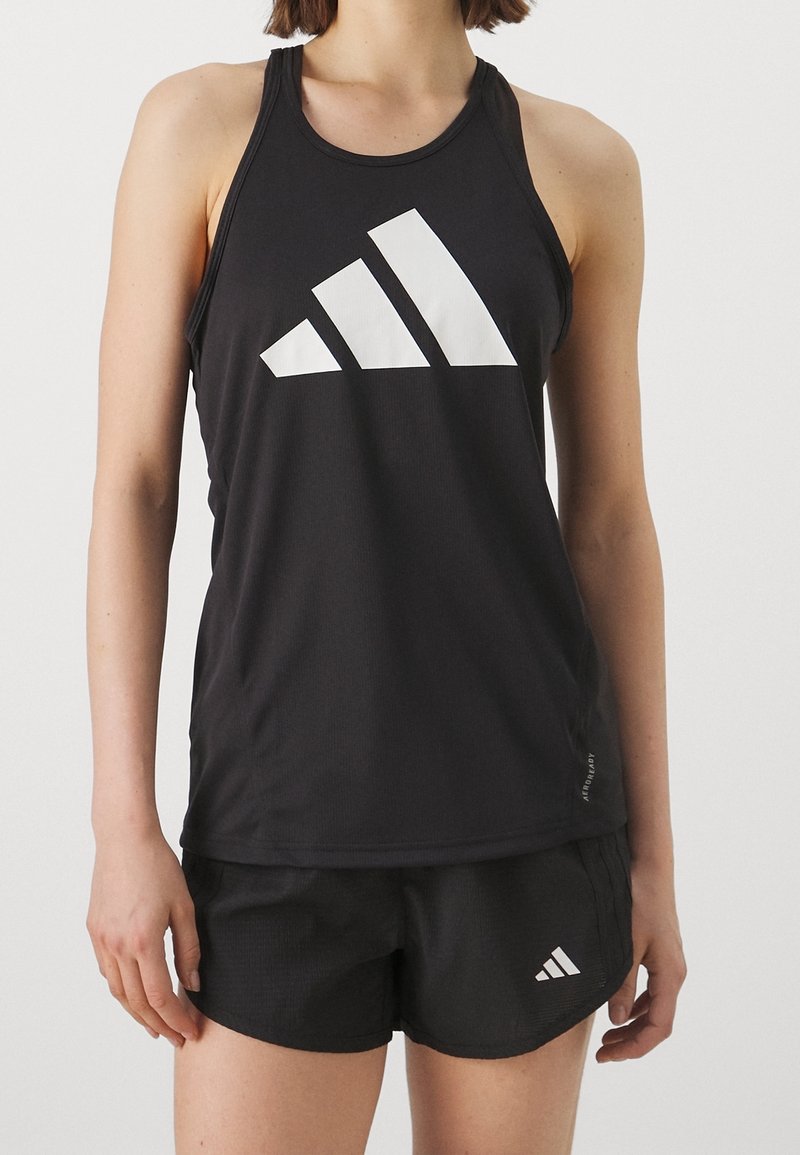 Femme portant un haut de sport noir sans manches et un short assorti avec des logos Adidas blancs à l'avant.