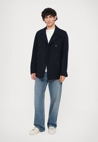 Abrigo de lana de mezcla en navy con diseño de doble botonadura, botones grandes y un cuello ancho, combinado con jeans de pierna ancha de color azul claro y zapatillas blancas.