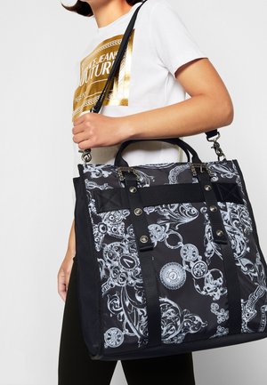 Persoon draagt een wit Versace Jeans Couture T-shirt en draagt een grote zwart-wit gevlekte boodschappentas met riemen en metalen details.