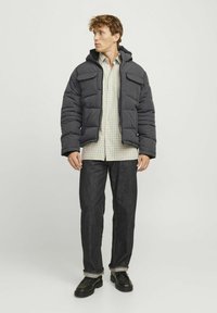Mørkegrå pufferjakke med hætte, med quiltede sektioner og fire frontlommer, båret over en lysternet skjorte og mørke jeans.