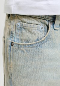 Φωτεινές μπλε τζιν παντελόνια με ορατή μπροστινή τσέπη και καρφιά, συνδυασμένα με λευκή μπλούζα με τελείωμα πάνω από τη μέση.