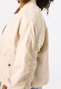 Beżowa kurtka bomberka o gładkiej, błyszczącej fakturze, z zamkiem błyskawicznym, prążkowanym kołnierzem i elastycznymi rękawami. Posiada boczne kieszenie i logo.