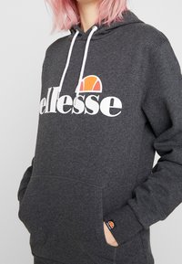 Sudadera gris con logo blanco de "ellesse" y acento naranja. Presenta un bolsillo frontal, capucha con cordón y puños acanalados. Textura suave.
