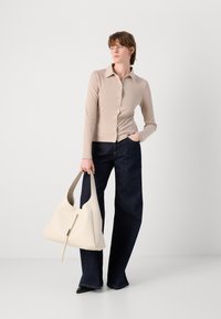 Beige gerippte Knopf-Oberteil, dunkelblaue weite Jeans und eine cremefarbene Hobo-Tasche mit einem Reißverschlussdetails. Das Model steht mit einer Hand in der Tasche.