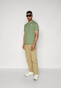 Camiseta de algodón verde con un pequeño logo, combinada con pantalones cargo de color beige claro con cremalleras laterales y zapatillas deportivas blancas.