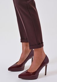 Tacchi alti in pelle patinata bordeaux con punta affilata, caratterizzati da un design elegante e un tacco a stiletto alto. I pantaloni sono arrotolati ai polsini.