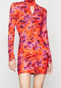 Robe mini à manches longues en tissu orange vif avec des motifs floraux violets et roses, col haut avec une ouverture en forme de clé, et des détails froncés.