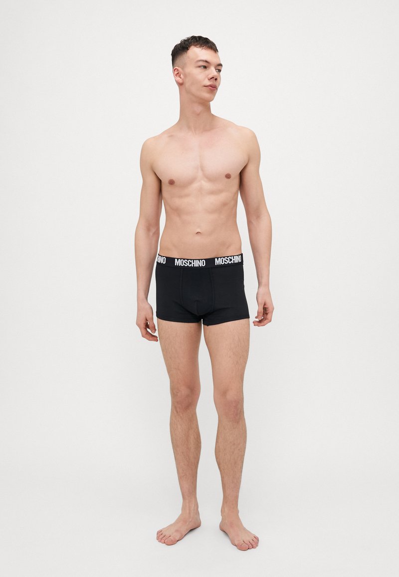 Black cotton boxer shorts with a white Moschino logo waistband, featuring a snug fit and minimal seam detailing. Model stands barefoot.