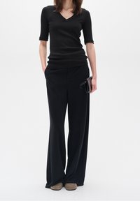 Svart ribbad topp med V-hals och korta ärmar, åtföljd av lösa svarta byxor. Modellen håller en liten svart kuvertväska och bär beige skor.