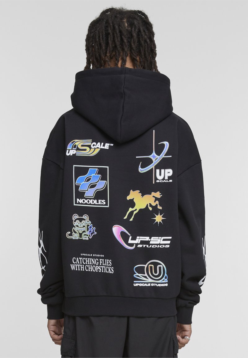 Persoon met dreadlocks draagt een zwarte hoodie met verschillende kleurrijke logo's, symbolen en teksten waaronder "NOODLES" en "VANGEN VAN VLIEGEN MET ETENSTOKJES."