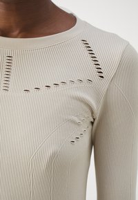 Närbild av en beige ribbad stickad topp med dekorativa utskärningsmönster runt bröstet och axelområdet.