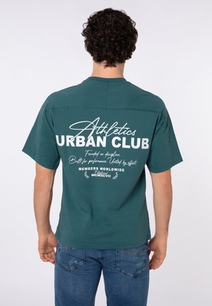 Mann mit lockigem Haar, der ein türkisfarbenes "Athletics Urban Club" T-Shirt und blaue Jeans trägt, steht mit dem Rücken zur Kamera.