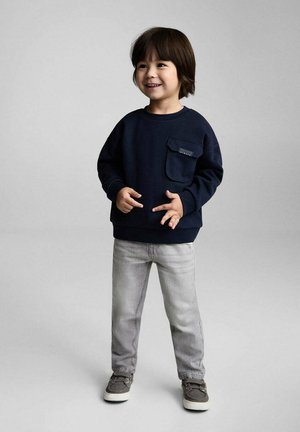 Blå sweatshirt med en frontlomme, parret med lysegrå tapered jeans og grå sko. Barnet står med hænderne foran og smiler.