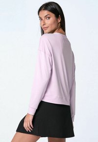 Donna con lunghi capelli scuri che indossa una maglietta a maniche lunghe viola chiaro e una gonna nera, che guarda oltre la spalla su uno sfondo bianco.