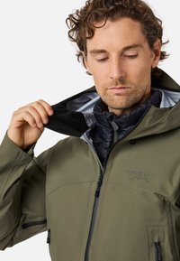Giacca impermeabile verde oliva con cappuccio, dotata di chiusura frontale con zip, finitura strutturata e logo sul petto. Strato interno nero visibile.
