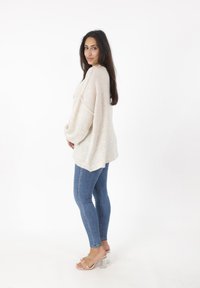 Maglione oversize beige chiaro con spalle basse, abbinato a jeans skinny blu e sandali con tacco trasparente. Texture semplice e morbida.