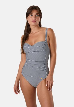 Donna che indossa un costume intero a righe bianche e nere con dettaglio arricciato davanti e spallina singola, in piedi su sfondo bianco.