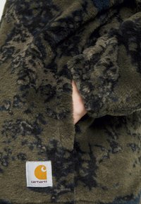Fleecejacke in Dunkelgrün mit schwarzen Mustern, zu sehen ist eine Nahaufnahme einer Hand in der Tasche und ein sichtbares Carhartt Logo-Patch.