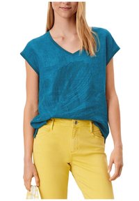 Mujer vestida con una blusa de manga corta en azul verdoso y escote en V con un patrón de hojas, metida dentro de unos pantalones amarillos brillantes, sosteniendo una tela con la mano izquierda.