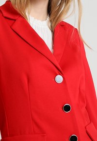 Blazer rouge avec une coupe ajustée, doté d'un revers à crans, de trois grands boutons noirs et d'une texture de tissu lisse.
