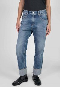 Persona que lleva puestos unos vaqueros de denim azul remangados, mocasines negros y calcetines grises, de pie contra un fondo claro.