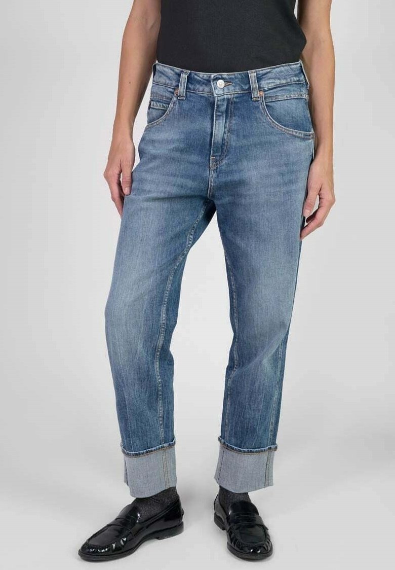 Persona que lleva puestos unos vaqueros de denim azul remangados, mocasines negros y calcetines grises, de pie contra un fondo claro.