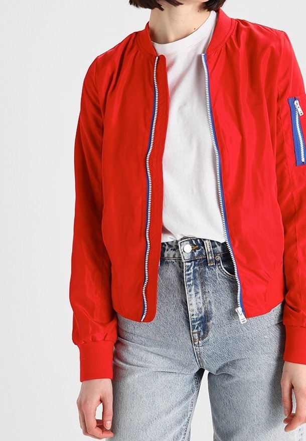 Veste bombers rouge en matière légère, avec une poche zippée bleue, des poignets côtelés et une fermeture zippée simple à l'avant. Portée avec un jean en denim.