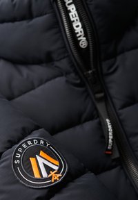 Marineblauwe pufferjas met een geborduurde textuur, voorzien van een Superdry-logo patch in zwart, oranje en wit; inclusief een zwarte rits.