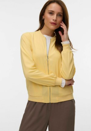 Žena s hnedými vlasmi, ktorá nosí svetložltú vetrovku na zips cez bielu košeľu a hnedé nohavice, drží jednu ruku blízko tváre.