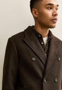 Abrigo de mezcla de lana marrón, de doble botonadura, con solapas de pico, que presenta botones oscuros y una camisa marrón texturizada debajo.