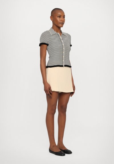 Camiseta de botones a rayas en blanco y negro con mangas cortas y bordes rizados, combinada con una mini falda beige y zapatos planos negros.