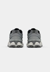 Ein Paar graue und schwarze New Balance Sneaker von hinten auf einem weißen Hintergrund mit dicke weißen und schwarzen Sohlen gezeigt.
