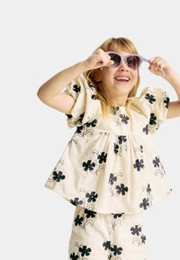 Haut en coton beige avec des manches bouffantes, orné d'un motif floral noir. Enfant portant des lunettes de soleil lilas, souriant et ajustant ses lunettes.