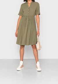 Vestido caqui de botonadura con mangas cortas, cinturilla fruncida y detalle de lazo en el frente, combinado con zapatillas blancas y beige y un bolso pequeño.