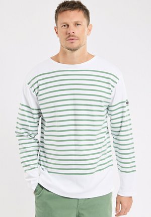 Homme portant une chemise blanche à manches longues avec des rayures horizontales vertes et un pantalon vert clair, debout devant un fond uni.