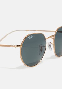 Kulta-toninen metalliset aurinkolasit pyöreillä tummilla linsseillä. Ominaisuuksia ovat langasta valmistettu kehys, teksturoitu koriste, ja nenätyynyt mukavuutta varten. Ray-Ban-logo näkyvissä.