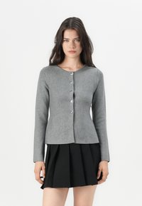 Grå strikkegenser med en tettsittende design, rund halslinning og fem dekorative knapper foran. Brukt over et plisserte svart skjørt.