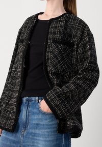 Femme portant une veste texturée à carreaux noir et blanc par-dessus un haut noir et un jean bleu, une main dans la poche, sur un fond uni.
