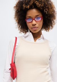 Γυναίκα με κατσαρά μαλλιά που φοράει κόκκινα γυαλιά, μπεζ τοπ πάνω από λευκό πουκάμισο και κρατάει κόκκινη τσάντα ώμου μπροστά σε απλό φόντο.