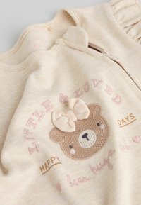 Mono beige para toddler con cierre de cremallera, que presenta un diseño bordado de un oso, corazones y el texto "Pequeño y Querido" en colores pastel. Textura suave.