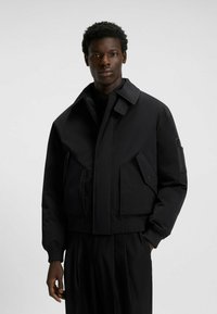 Sort bomberjakke med en struktureret design, der har store frontlommer og en lynlåslomme på ærmet. Glat tekstur og cropped pasform.