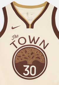 Kremfarget basketballtrøye med brune detaljer. Har V-hals, teksten "The Town", en tre-grafikk og tallet "30" i hvitt.