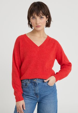 Donna con capelli corti castani che indossa orecchini a cerchio argentati, maglione rosso con scollo a V e jeans a vita alta blu con una mano in tasca.