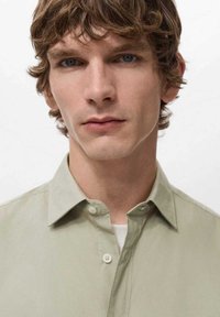 Camisa de manga larga de color verde claro con cuello clásico y textura suave, llevada sobre una camiseta interior blanca. Expresión facial clara.