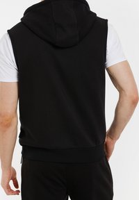 Hoodie noir sans manches en tissu doux ; doté d'une capuche spacieuse, d'un ourlet côtelé et de fentes latérales pour le confort et la liberté de mouvement.