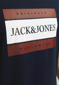 Marinblå t-shirt med en stor rektangulär grafik med vit mitt och bruna accentfärger, som visar "JACK&JONES" och "ORIGINALS WORLDWIDE."