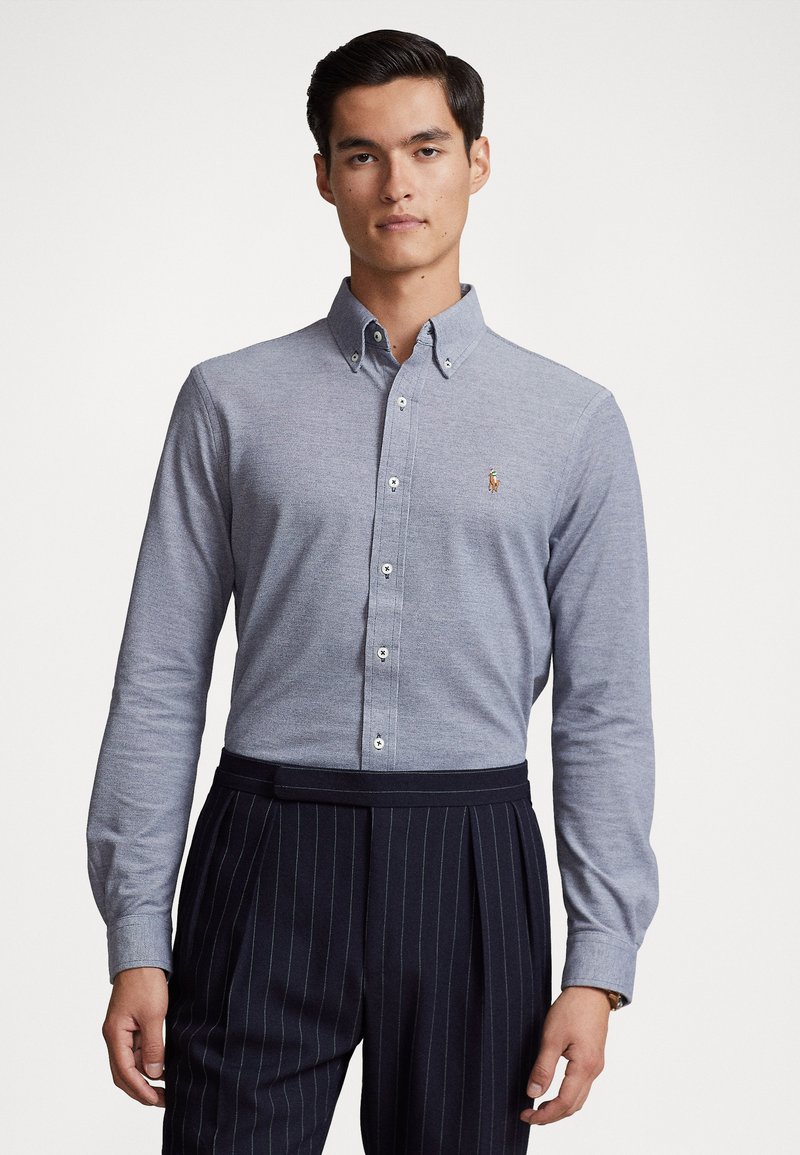 Lange mouw knoopshirt van lichtgrijze stof, met een klein geborduurd logo, gecombineerd met marineblauwe broek met streepjes.