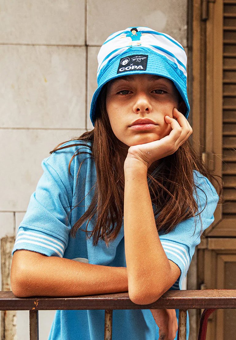 Blauwe bucket hat met witte strepen en logo-tag, gecombineerd met een casual blauwe shirt. Handen rusten op een metalen reling. Natuurlijke haartextuur.